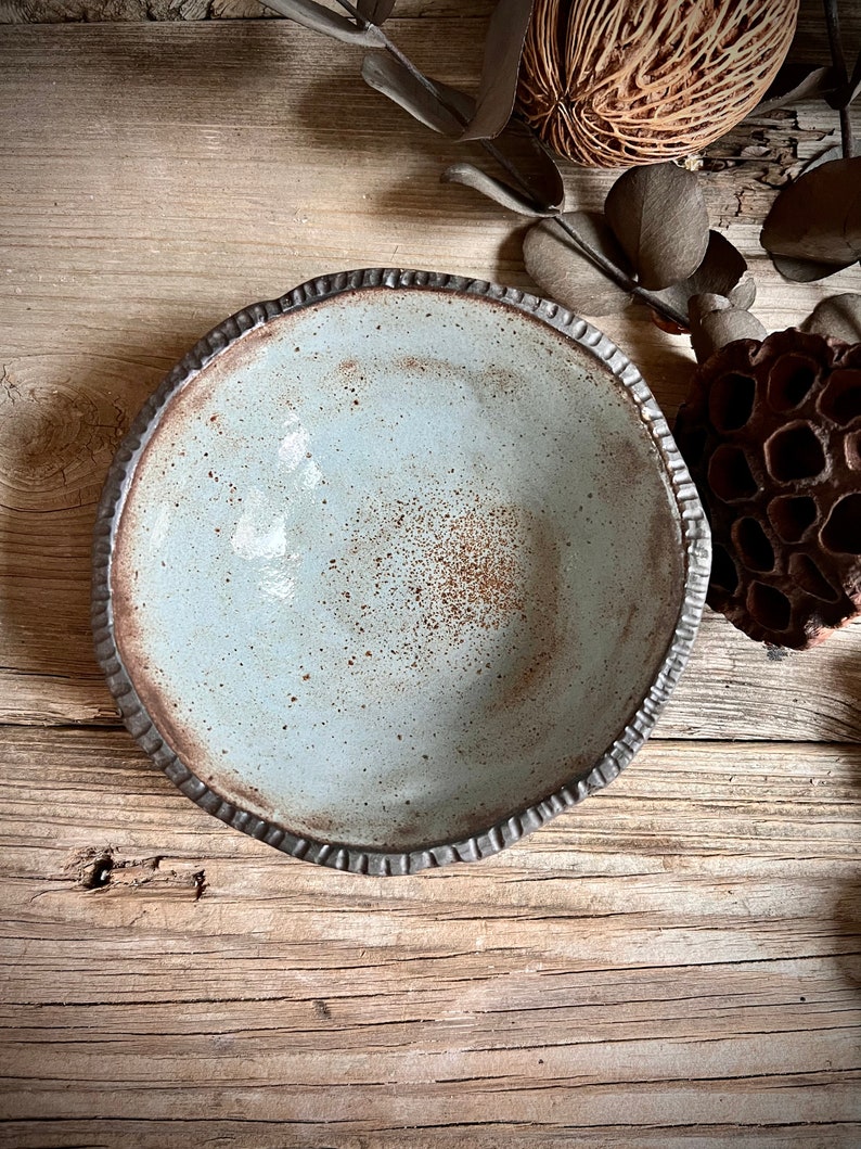 May include: A light blue ceramic bowl with a textured rim. The bowl has a speckled brown glaze and is sitting on a wooden surface.