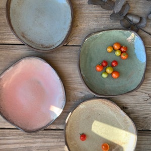 May include: Four ceramic plates in different colors and shapes. The plates are on a wooden surface. One plate has small red tomatoes on it.