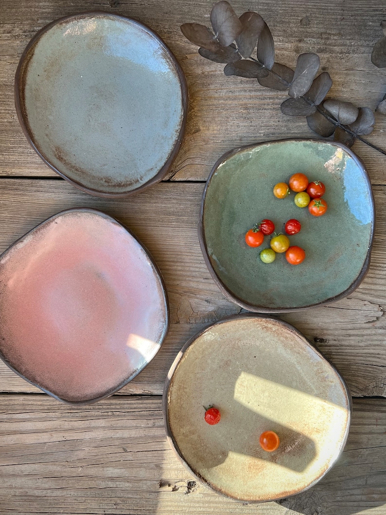May include: Four ceramic plates in different colors and shapes. The plates are on a wooden surface. One plate has small red tomatoes on it.