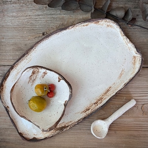 May include: A handmade ceramic serving set featuring a large rectangular platter, a smaller irregular-shaped bowl, and a spoon. The set is off-white with brown edges and speckles. The bowl contains yellow and red fruit. The set is on a wooden surface.