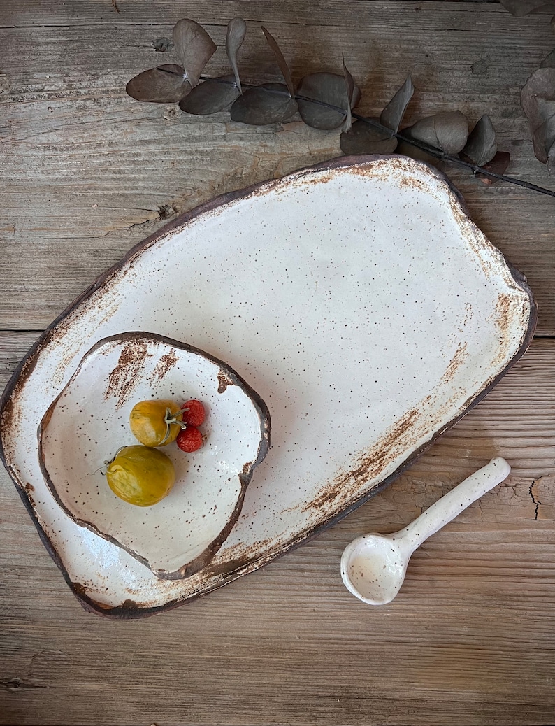 May include: A handmade ceramic serving set featuring a large rectangular platter, a smaller irregular-shaped bowl, and a spoon. The set is off-white with brown edges and speckles. The bowl contains yellow and red fruit. The set is on a wooden surface.