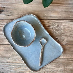 May include: A handmade ceramic serving set in a light blue speckled glaze. The set includes a rectangular tray, a small bowl, and a spoon. The edges have a contrasting brown trim. The set is displayed on a wooden surface.