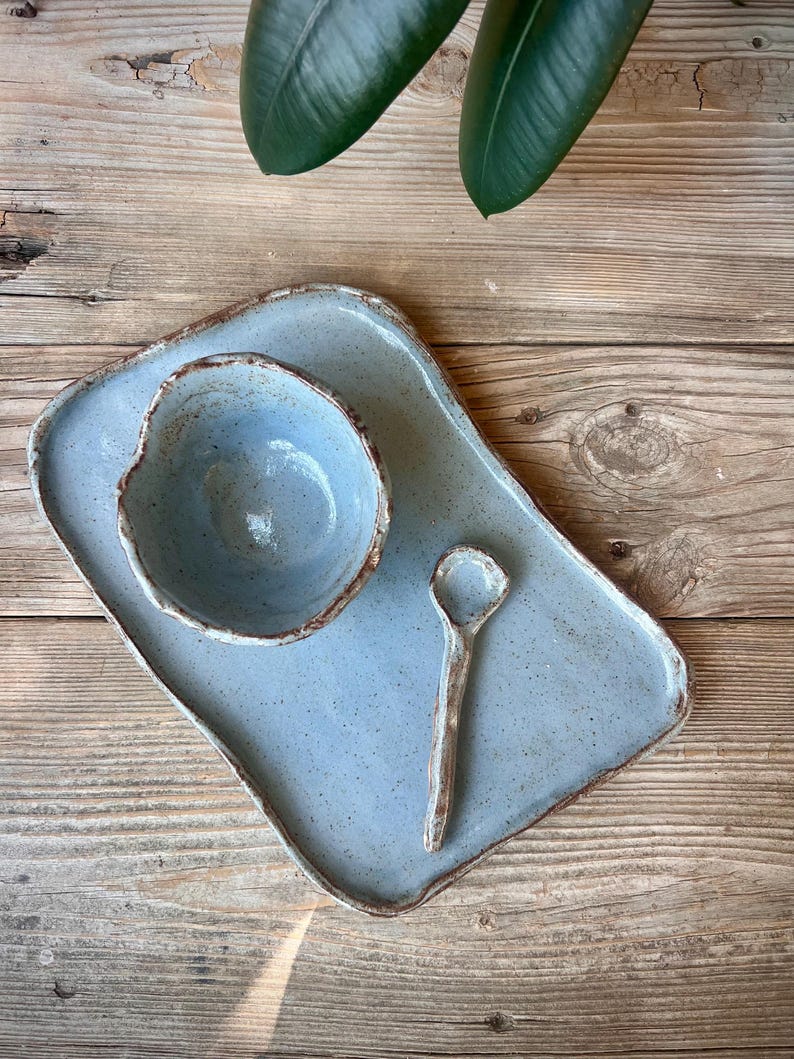 May include: A handmade ceramic serving set in a light blue speckled glaze. The set includes a rectangular tray, a small bowl, and a spoon. The edges have a contrasting brown trim. The set is displayed on a wooden surface.