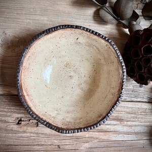 May include: A white ceramic bowl with a brown rim and a speckled glaze. The bowl is sitting on a wooden surface.