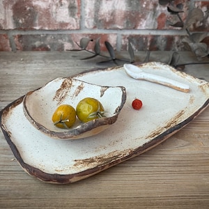 May include: A handmade ceramic serving set with a rectangular platter, a smaller bowl, and a spoon. The set is off-white with brown edges and speckles. Two yellow tomatoes and one red tomato are in the bowl and on the platter.