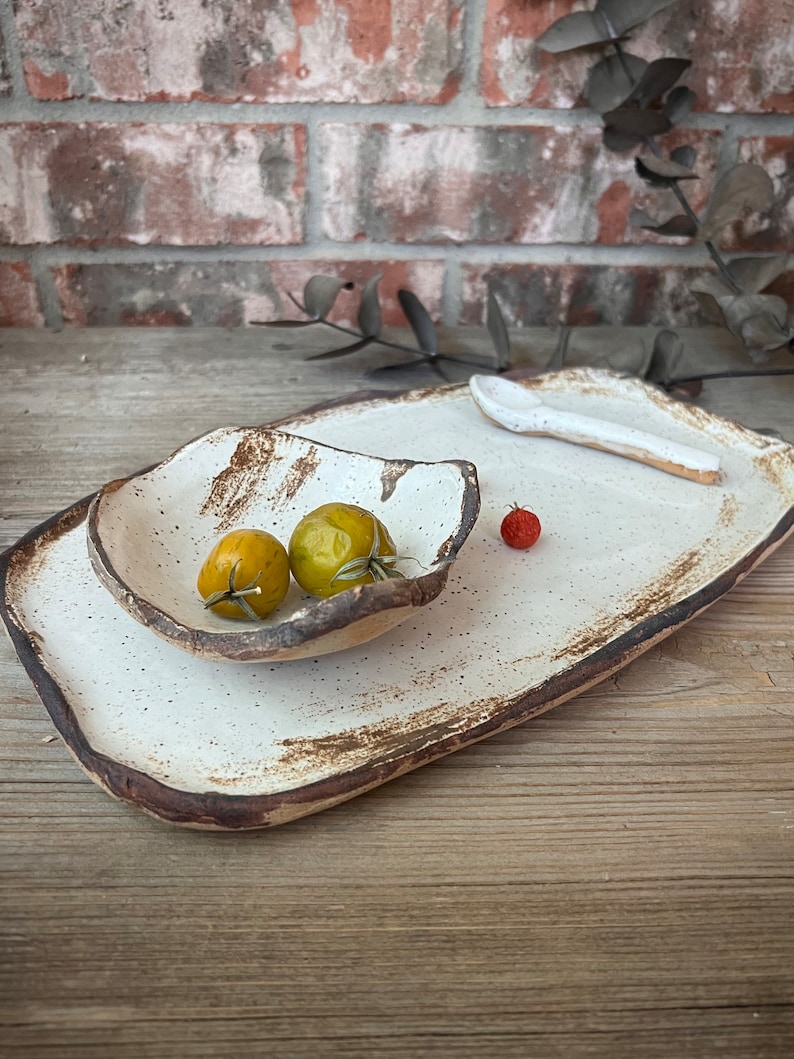 May include: A handmade ceramic serving set with a rectangular platter, a smaller bowl, and a spoon. The set is off-white with brown edges and speckles. Two yellow tomatoes and one red tomato are in the bowl and on the platter.