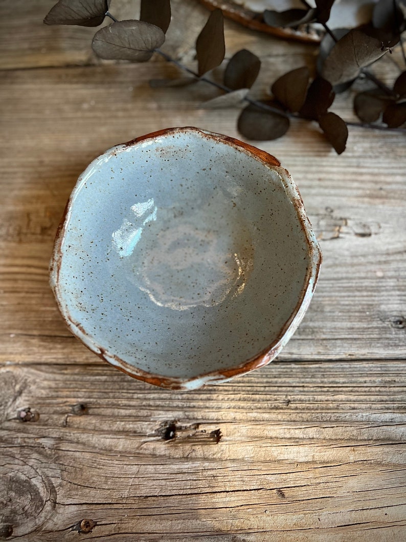 May include: A small, light blue ceramic bowl with a speckled glaze and a brown rim. The bowl is sitting on a wooden surface.