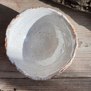 Puede incluir: Un cuenco de cer&aacute;mica blanca con un esmalte moteado y un borde ligeramente irregular. El cuenco est&aacute; sobre una superficie de madera.