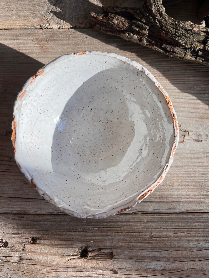 Puede incluir: Un cuenco de cer&aacute;mica blanca con un esmalte moteado y un borde ligeramente irregular. El cuenco est&aacute; sobre una superficie de madera.