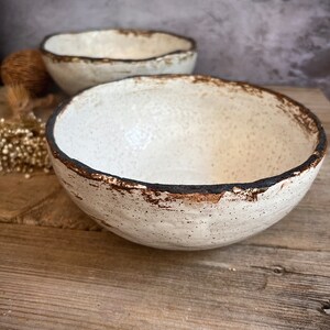May include: Two white ceramic bowls with a speckled glaze and a dark brown rim. The bowls are sitting on a wooden surface.