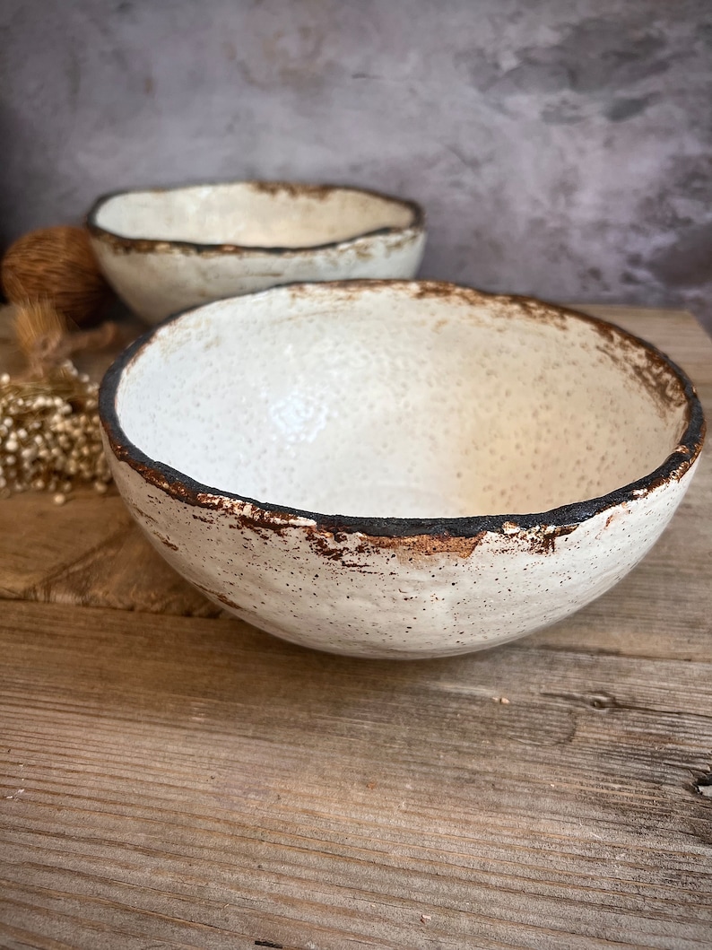 May include: Two white ceramic bowls with a speckled glaze and a dark brown rim. The bowls are sitting on a wooden surface.