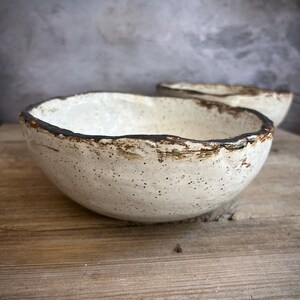 May include: A white ceramic bowl with a speckled glaze and a dark brown rim. The bowl is sitting on a wooden surface.