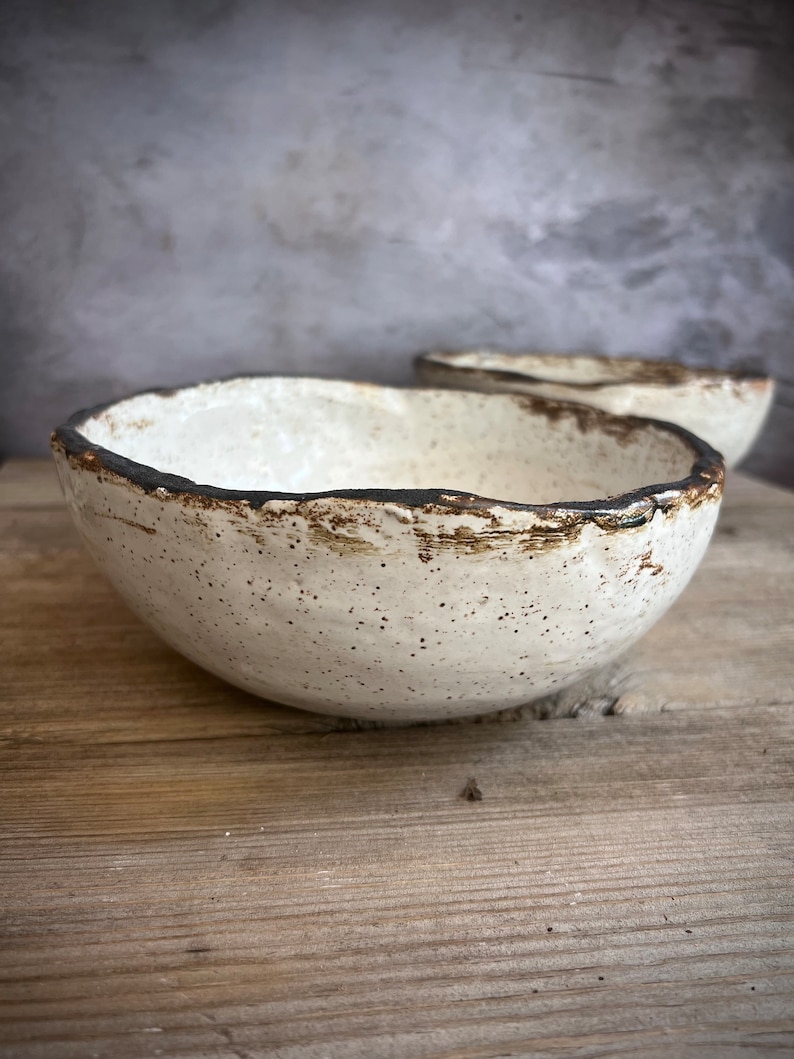 May include: A white ceramic bowl with a speckled glaze and a dark brown rim. The bowl is sitting on a wooden surface.