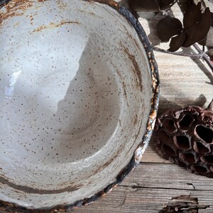 May include: A handmade ceramic bowl with a speckled white interior and a dark, textured rim. The bowl has a rustic, organic aesthetic, with visible imperfections and a natural color palette. The rim has a dark brown and black color. The bowl is likely used for serving or display.