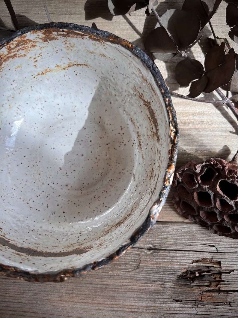 May include: A handmade ceramic bowl with a speckled white interior and a dark, textured rim. The bowl has a rustic, organic aesthetic, with visible imperfections and a natural color palette. The rim has a dark brown and black color. The bowl is likely used for serving or display.