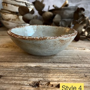 May include: A small, light blue ceramic bowl with a brown rim and speckled glaze. The bowl is sitting on a wooden surface. Style 4.