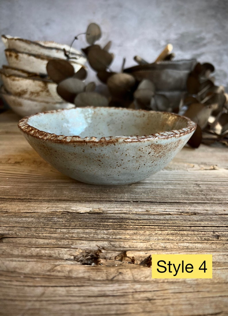 May include: A small, light blue ceramic bowl with a brown rim and speckled glaze. The bowl is sitting on a wooden surface. Style 4.