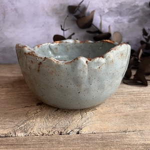 May include: A handmade, light blue ceramic bowl with a scalloped rim and speckled texture. The rim has a brown, rustic edge. The bowl is set on a wooden surface, showcasing its artisanal quality. This unique bowl is perfect for serving or display.
