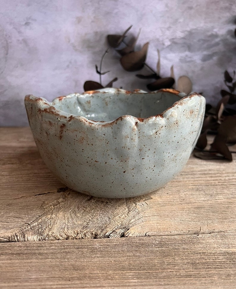 May include: A handmade, light blue ceramic bowl with a scalloped rim and speckled texture. The rim has a brown, rustic edge. The bowl is set on a wooden surface, showcasing its artisanal quality. This unique bowl is perfect for serving or display.