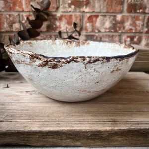 May include: A handcrafted white ceramic bowl with a textured surface and a rustic, uneven rim. The rim features a dark brown, speckled edge. The bowl is set on a weathered wooden surface against a brick background. This bowl is ideal for serving food.