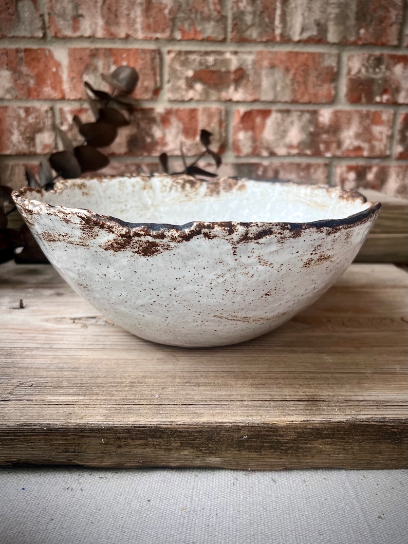 May include: A handcrafted white ceramic bowl with a textured surface and a rustic, uneven rim. The rim features a dark brown, speckled edge. The bowl is set on a weathered wooden surface against a brick background. This bowl is ideal for serving food.