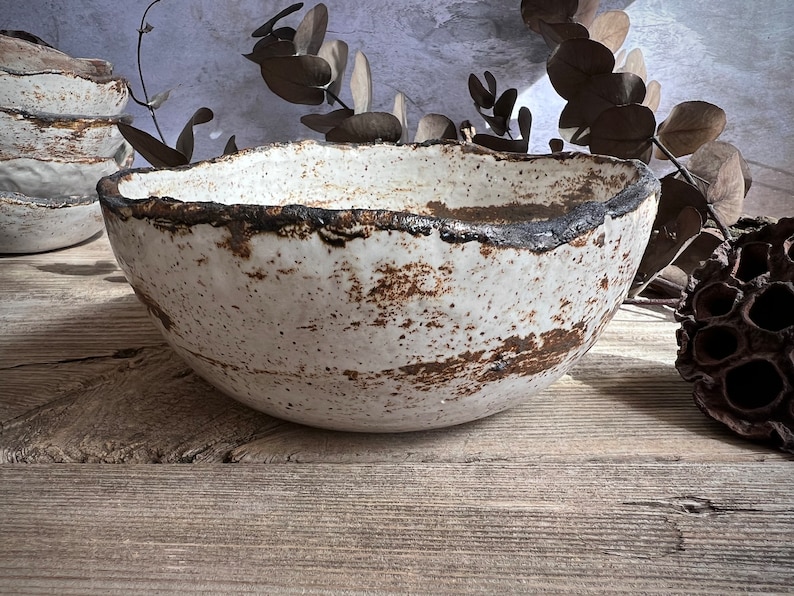 May include: A handmade white ceramic bowl with a textured surface and a dark brown rim. The bowl has a rustic, organic shape and is displayed on a wooden surface. Other bowls are visible in the background.