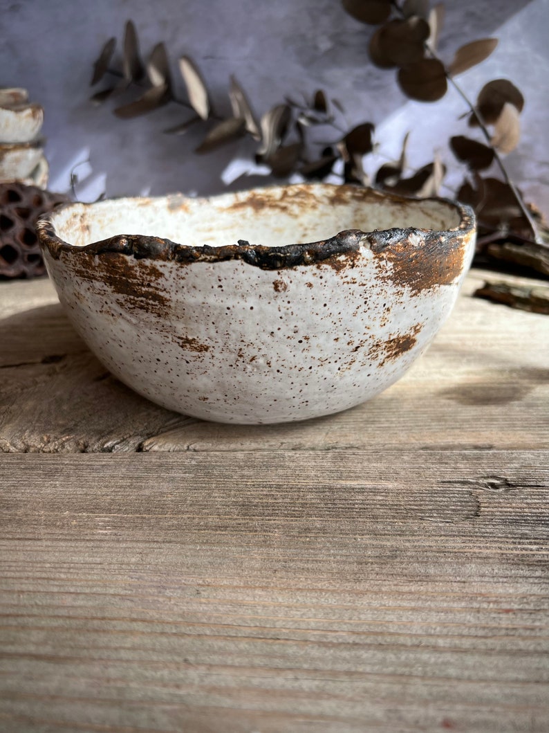 May include: A handmade ceramic bowl with a textured white exterior and a dark brown, uneven rim. The bowl has a rustic, organic aesthetic, perfect for serving food or as a decorative piece. The bowl sits on a weathered wooden surface.