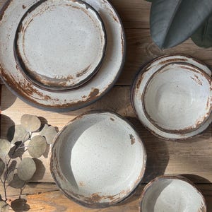 May include: A collection of handmade ceramic plates and bowls. The tableware features a speckled, off-white glaze with a rustic, brown rim. The plates and bowls vary in size and shape, creating a unique, artisanal aesthetic. The set is arranged on a wooden surface.