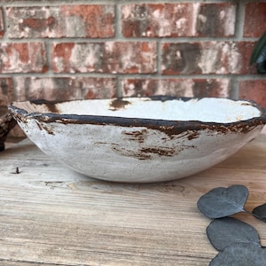 May include: A handmade, white ceramic bowl with a textured surface and a dark brown rim. The bowl has a rustic, organic shape and is displayed on a wooden surface. The background features a brick wall.