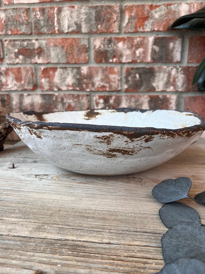 May include: A handmade, white ceramic bowl with a textured surface and a dark brown rim. The bowl has a rustic, organic shape and is displayed on a wooden surface. The background features a brick wall.