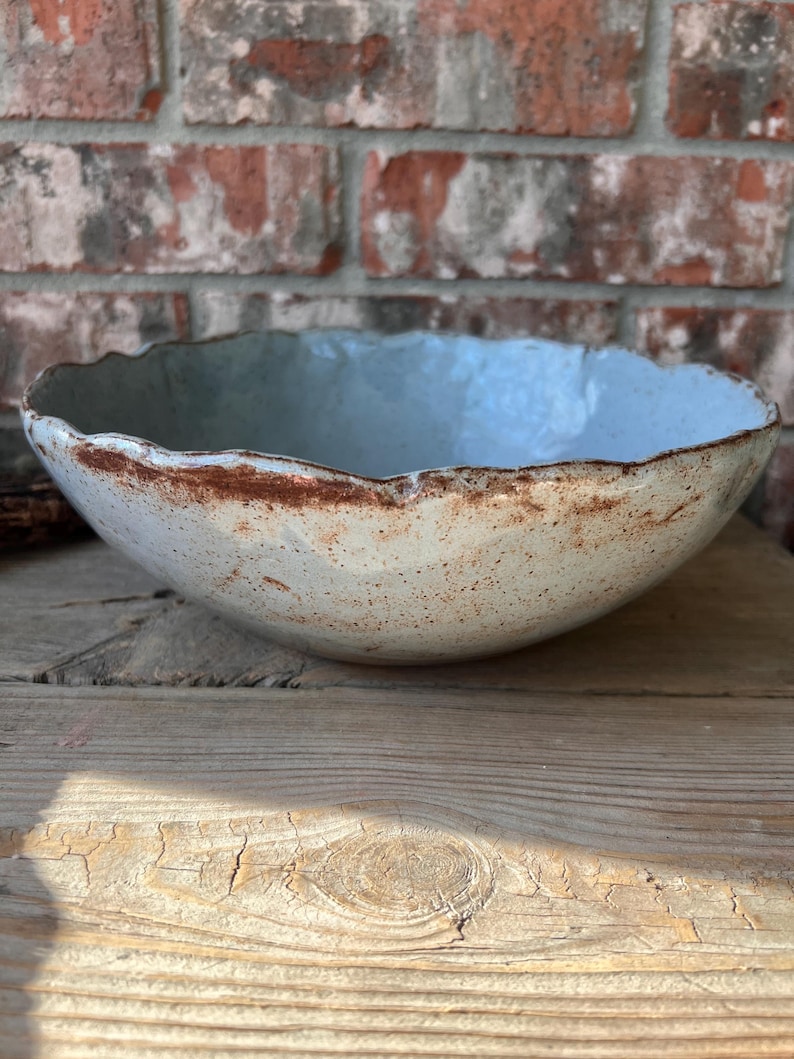 May include: A handmade ceramic bowl with a light blue interior and a textured, off-white exterior. The rim has a rustic, brown edge. The bowl sits on a wooden surface, with a brick wall in the background.