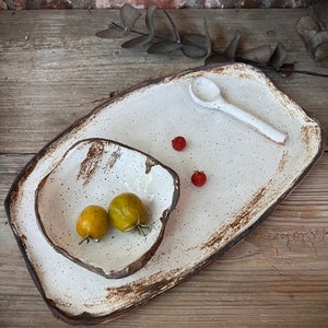 May include: A handmade ceramic serving set featuring a large rectangular platter, a smaller square bowl, and a spoon. The set has a speckled white glaze with a dark brown rim. Two yellow-green cherry tomatoes and two red berries are on the set.