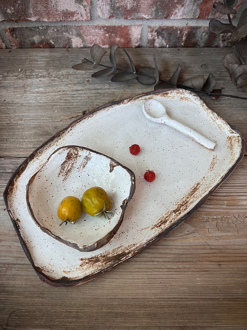 May include: A handmade ceramic serving set featuring a large rectangular platter, a smaller square bowl, and a spoon. The set has a speckled white glaze with a dark brown rim. Two yellow-green cherry tomatoes and two red berries are on the set.