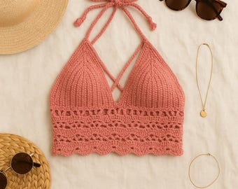 May include: A coral-colored, handmade crochet halter top with a crisscross design. The top features a scalloped edge and is displayed with sunglasses, a necklace, and a straw hat, suggesting a summer theme.