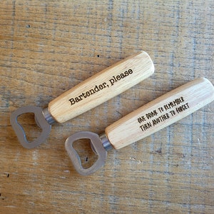 May include: Two wooden bottle openers with stainless steel heads. One opener reads "Bartender, please" and the other reads "ONE DRINK TO REMEMBER THEN ANOTHER TO FORGET". The openers are on a light brown wooden surface.