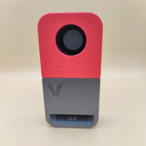 Op de afbeelding: Een tweekleurig apparaat met een rood bovenstuk en een grijs onderstuk. Het rode gedeelte heeft een cirkelvormige opening en de tekst "Bento mini". Het grijze gedeelte heeft een gestileerd "V"-logo.