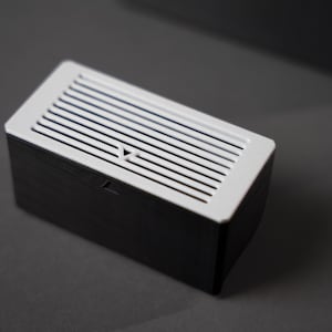 K&ouml;nnte beinhalten: Eine rechteckige, schwarz-wei&szlig;e Box mit einem Lattenrost. Die Oberseite ist wei&szlig; mit parallelen Schlitzen und der K&ouml;rper ist schwarz. Die Box hat einen kleinen, dreieckigen Ausschnitt auf der Vorderseite.
