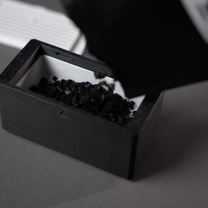 K&ouml;nnte beinhalten: Eine schwarze Holzkiste mit wei&szlig;em Innenraum wird mit kleinem, dunklem, k&ouml;rnigem Material gef&uuml;llt. Die Kiste ist rechteckig und steht auf einer grauen Oberfl&auml;che. Ein wei&szlig;es, lamellenartiges Objekt befindet sich im Hintergrund.