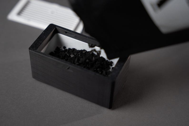 K&ouml;nnte beinhalten: Eine schwarze Holzkiste mit wei&szlig;em Innenraum wird mit kleinem, dunklem, k&ouml;rnigem Material gef&uuml;llt. Die Kiste ist rechteckig und steht auf einer grauen Oberfl&auml;che. Ein wei&szlig;es, lamellenartiges Objekt befindet sich im Hintergrund.