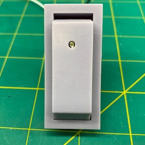 Puede incluir: Un interruptor de palanca gris con un pequeño botón redondo blanco en el centro. El interruptor está montado en una base de plástico blanco.