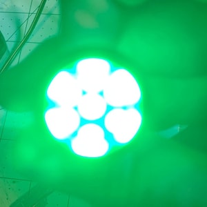 Puede incluir: Una luz LED verde con forma circular y múltiples luces pequeñas dispuestas en forma de flor. La luz está siendo sostenida por una mano.