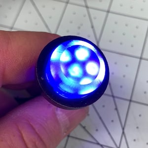 Puede incluir: Primer plano de una pequeña luz redonda negra con una luz LED azul en su interior. La luz está encendida e ilumina el área circundante.