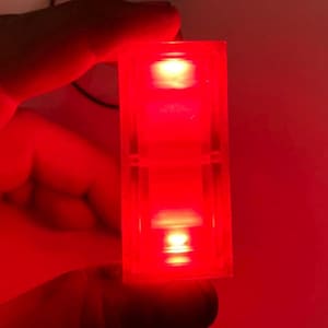 Puede incluir: Un bloque de plástico rojo con dos luces LED rojas en su interior. Las luces están encendidas y brillan.