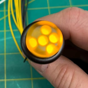 Puede incluir: Primer plano de una pequeña luz LED redonda amarilla con cinco LED individuales. La luz está conectada a cables negros.