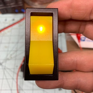 Puede incluir: Un interruptor de palanca amarillo con un marco negro. El interruptor está iluminado con una luz naranja brillante.