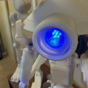 Könnte beinhalten: Ein weißer, beweglicher Roboter mit einem großen, kreisförmigen Auge, das ein hellblaues Licht aussendet. Der Roboter hat mehrere Gelenke und einen kastenförmigen Körper, was auf ein mechanisches oder futuristisches Thema hindeutet. Die komplizierte Innenstruktur des Auges ist sichtbar.