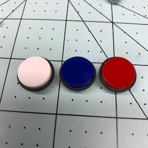 Puede incluir: Tres botones redondos con bordes negros. Los botones son blancos, azules y rojos.