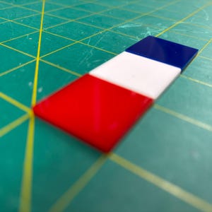 May include: A rectangular object with three colored sections: red, white, and blue. The object is positioned diagonally on a green cutting mat with yellow grid lines. The colors are vibrant and distinct.