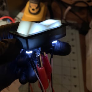 Puede incluir: Un interruptor rectangular negro con una superficie blanca iluminada. El interruptor está cableado con cables rojos y blancos.