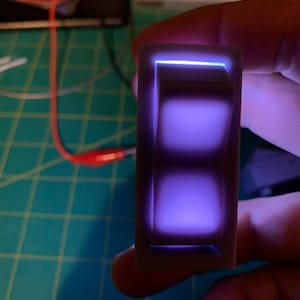 Puede incluir: Un interruptor de palanca de plástico blanco con un brillo púrpura que emana del interior. El interruptor está en la posición de apagado.